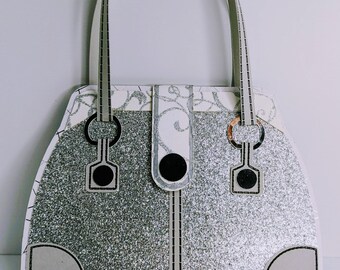 Op de afbeelding: Een zilveren glitterhandtas met een wit bloemenpatroon bovendeel en een bijpassende handgreep. De tas heeft zwarte accenten, waaronder een sluiting en decoratieve elementen. Het algehele ontwerp is symmetrisch en elegant.