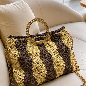 May include: A woven tote bag with a brown and tan striped pattern. The bag has a gold chain strap and a gold chain handle. The bag is sitting on a white couch with a view of the ocean in the background.