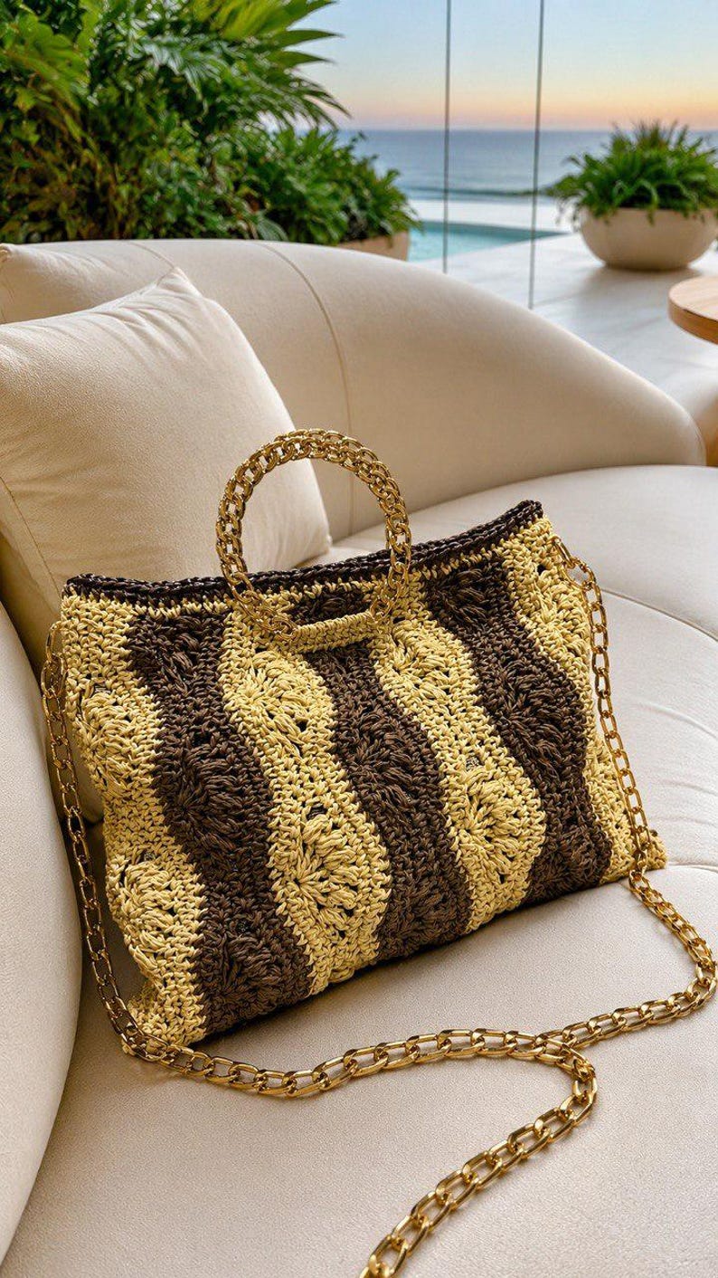 May include: A woven tote bag with a brown and tan striped pattern. The bag has a gold chain strap and a gold chain handle. The bag is sitting on a white couch with a view of the ocean in the background.