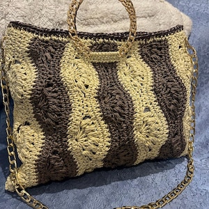 May include: A handmade crochet handbag with a gold chain handle and strap. The bag features a striped pattern in shades of brown and beige. The gold chain adds a touch of elegance to this stylish accessory.