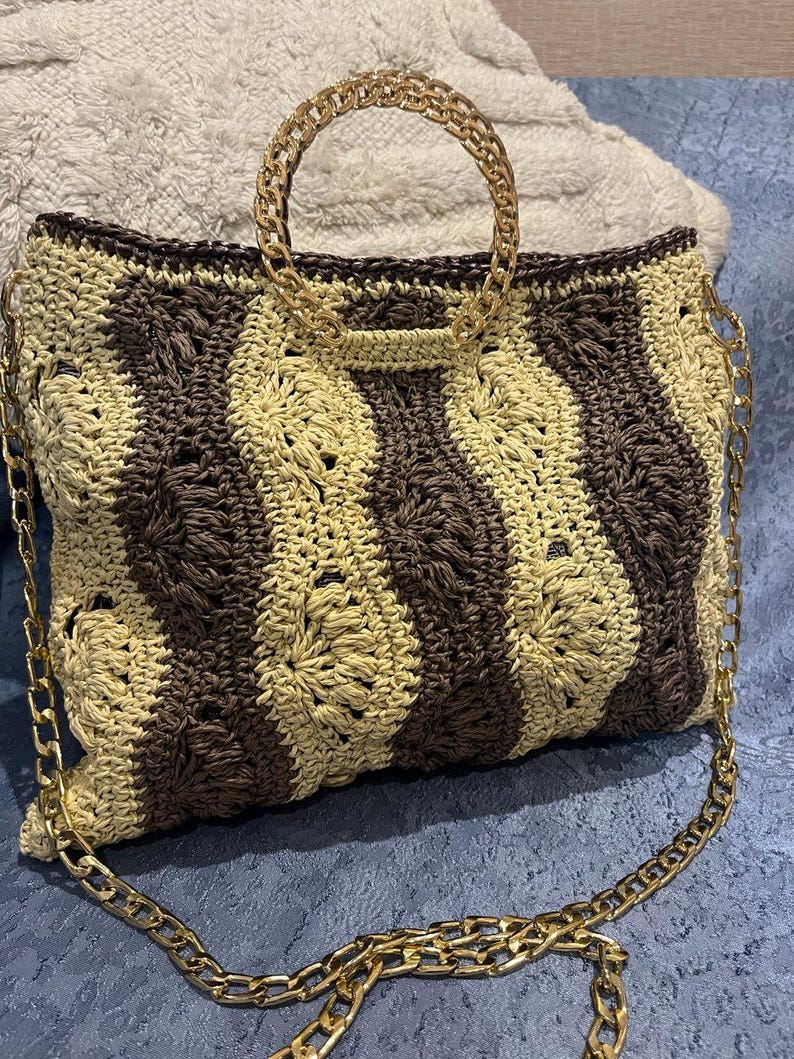 May include: A handmade crochet handbag with a gold chain handle and strap. The bag features a striped pattern in shades of brown and beige. The gold chain adds a touch of elegance to this stylish accessory.