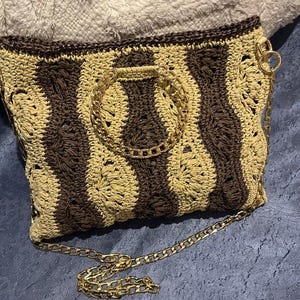 May include: A crocheted handbag with a brown and tan striped pattern. The bag features a gold chain strap and a gold chain handle. The bag is rectangular and has a dark brown trim.