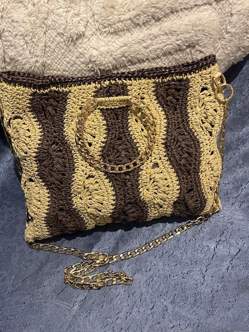 May include: A crocheted handbag with a brown and tan striped pattern. The bag features a gold chain strap and a gold chain handle. The bag is rectangular and has a dark brown trim.