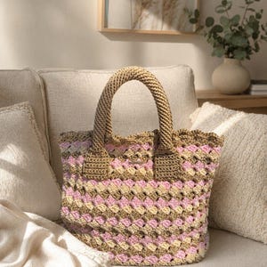 May include: A crochet tote bag featuring a repeating granny square pattern in alternating horizontal stripes of tan, light pink, and cream yarn. The bag has two thick, braided tan handles and a scalloped top edge with a small rectangular metal tag.