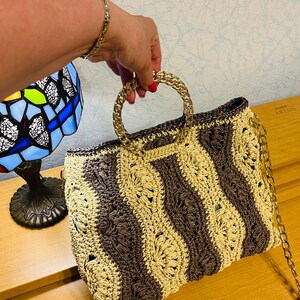 May include: A handmade crochet handbag with a gold chain strap and gold handles. The bag features a striped pattern in shades of brown and beige. The bag is held by a person.