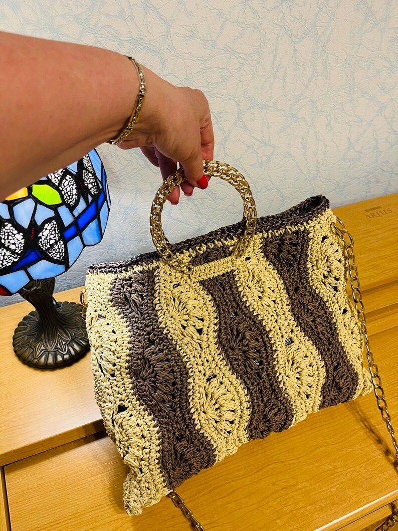 May include: A handmade crochet handbag with a gold chain strap and gold handles. The bag features a striped pattern in shades of brown and beige. The bag is held by a person.