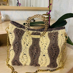 May include: A hand-crocheted handbag with a woven pattern in alternating vertical stripes of beige and brown. The bag features a gold-colored chain strap and a circular handle. The bag is sitting on a wooden surface.