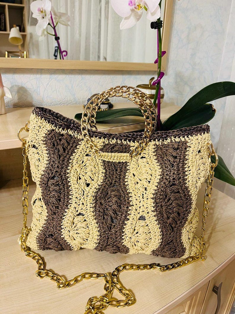 May include: A hand-crocheted handbag with a woven pattern in alternating vertical stripes of beige and brown. The bag features a gold-colored chain strap and a circular handle. The bag is sitting on a wooden surface.