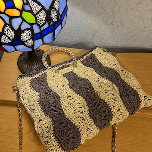 May include: A handmade crochet handbag with a wavy striped pattern in beige and dark gray. The bag features a gold chain strap and a clear handle. The bag is sitting on a wooden surface.