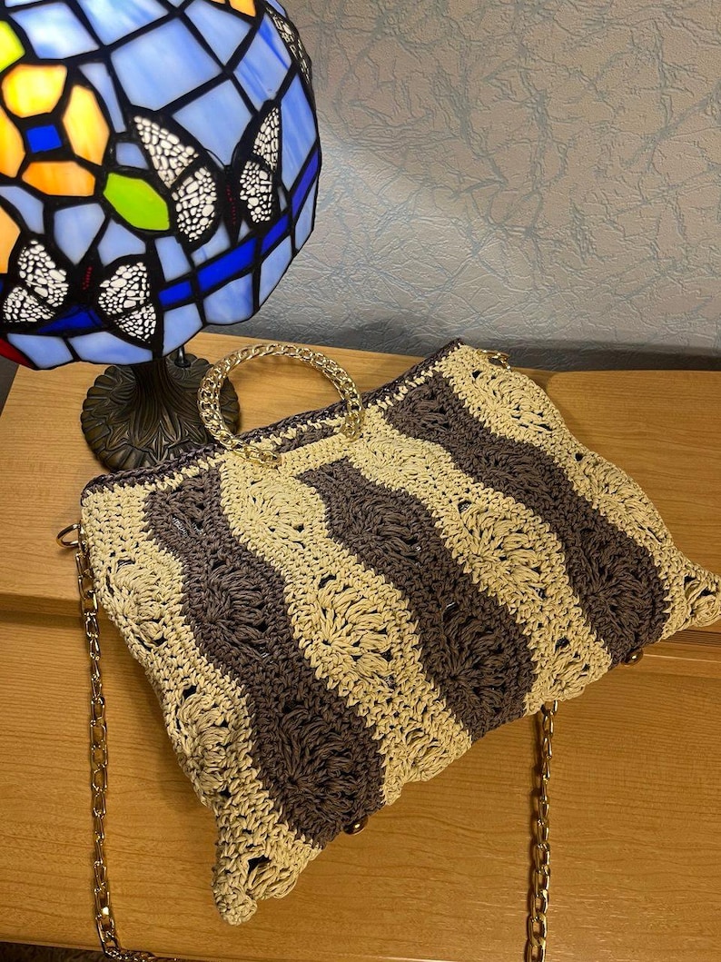 May include: A handmade crochet handbag with a wavy striped pattern in beige and dark gray. The bag features a gold chain strap and a clear handle. The bag is sitting on a wooden surface.