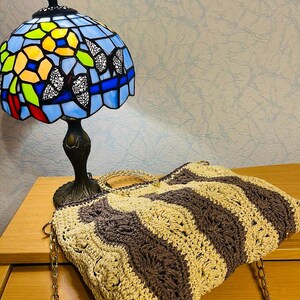 May include: A Tiffany-style table lamp with a blue stained glass shade featuring butterfly and floral designs. A handmade crochet tote bag with brown and beige wavy stripes and gold chain straps rests nearby.