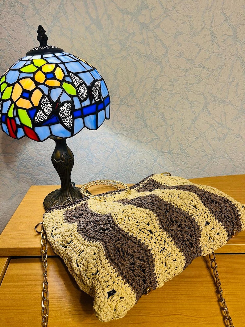 May include: A Tiffany-style table lamp with a blue stained glass shade featuring butterfly and floral designs. A handmade crochet tote bag with brown and beige wavy stripes and gold chain straps rests nearby.