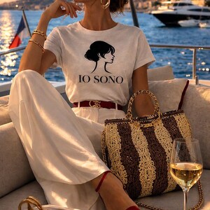 May include: A woman wearing a white t-shirt with a black graphic and the text "IO SONO", white pants, and red sandals. She is sitting on a boat with a striped handbag, a glass of wine, and a book with the word "PARIS".