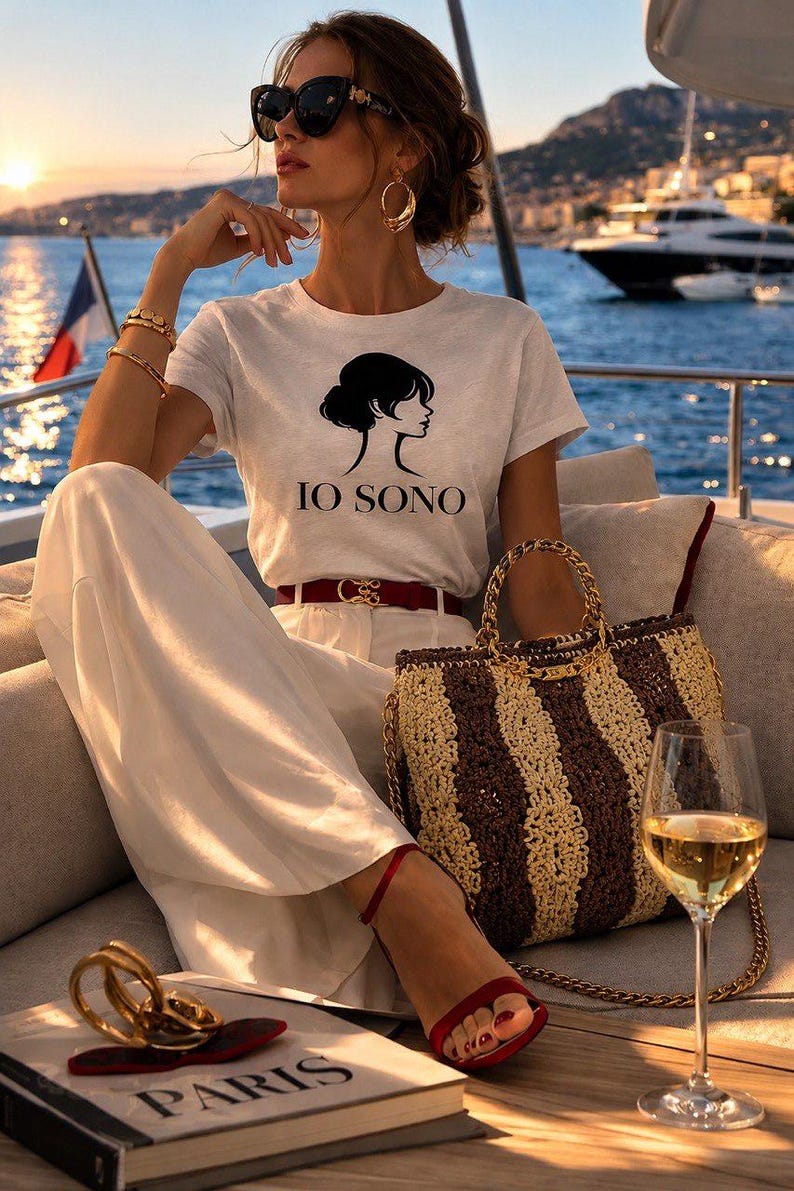 May include: A woman wearing a white t-shirt with a black graphic and the text "IO SONO", white pants, and red sandals. She is sitting on a boat with a striped handbag, a glass of wine, and a book with the word "PARIS".