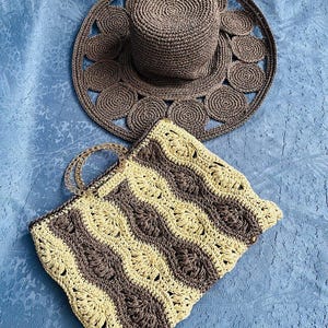 May include: A crocheted sun hat and handbag set. The hat is brown with a wide brim and circular accents. The handbag features a wavy pattern in cream and brown, with clear handles. The set is designed for summer.