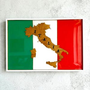 Könnte beinhalten: Ein gerahmtes Kunstwerk mit dem Design der italienischen Flagge. Die Flagge ist in grüne, weiße und rote vertikale Streifen unterteilt. Ein Holzausschnitt von Italien ist zentriert, mit dem Wort "ITALIA" darauf geschrieben. Das Kunstwerk ist in einem weißen Rahmen eingefasst.