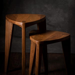 May include: Two wooden stools with triangular seats and angled legs. The stools are made of a warm-toned wood, with visible grain patterns. The background is a dark, textured gray.