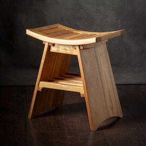 Könnte beinhalten: Ein Holzhocker mit geschwungener Sitzfläche und einer unteren Ablage aus Holzlatten. Der Hocker hat eine hellbraune Holzmaserung und ein Markenlogo auf der vorderen Stützstrebe. Das Design umfasst angewinkelte Seitenteile und eine Sitzfläche.