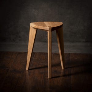 May include: A three-legged wooden stool with a triangular seat. The stool is made of light-colored wood and has a simple, modern design. The legs are angled and splay outward. The stool is shown against a dark background.