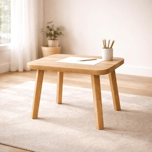 Op de afbeelding: Een kleine, vierkante houten tafel met afgeronde hoeken en vier stevige poten. Een vel papier met een potlood en een klein wit kopje met potloden staan erop. De tafel is gemaakt van lichtgekleurd hout.