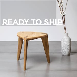 Peut inclure: Un petit tabouret en bois à trois pieds avec une assise triangulaire. Le tabouret est en bois clair et a une finition naturelle. Le texte "READY TO SHIP" est affiché en lettres blanches au-dessus du tabouret.