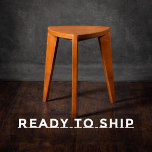 Może przedstawiać: Drewniany stołek z trójkątnym siedziskiem i trzema nogami. Stołek wykonany jest z jasnego drewna i ma prosty, minimalistyczny design. Tekst "READY_TO_SHIP" jest wyświetlany na dole obrazu na biało.