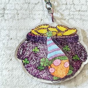 Puede incluir: Un llavero con temática del Día de San Patricio con una olla de oro, un duende y tréboles. El llavero tiene un cierre y una cadena de color arcoíris. La olla es morada y está llena de monedas de oro. El duende lleva un sombrero a rayas azules y blancas.