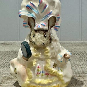 May include: A ceramic mouse figurine wearing a bonnet with pink and blue stripes, holding a small mirror. The mouse is dressed in a white dress with floral and yellow trim. The figurine is on a wooden surface.