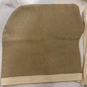 May include: A tan, textured fabric pouch with a rounded top corner and a straight bottom edge. The pouch features a cream-colored fabric trim along the bottom and side, extending into two long, flat fabric ties for securing the item closed.