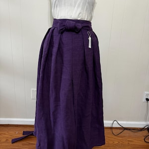 May include: A white sleeveless top paired with a long, deep purple skirt. The skirt features a bow at the waist and a flowing, pleated design. The outfit is displayed on a mannequin, showcasing the garment's shape and texture.