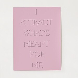 May include: A pale pink rectangular card featuring the embossed phrase "I ATTRACT WHAT'S MEANT FOR ME". The text is centrally aligned and in a sans-serif font. The card is set against a white backdrop.