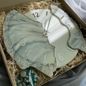 May include: A decorative wall clock with a white face and silver hands. The clock features a unique design with a marbled, light blue and white resin border. The numbers are silver. The clock is packaged in a box with decorative filler.