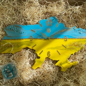 Puede incluir: Un reloj de pared con la forma del mapa de Ucrania, con una combinación de colores azul y amarillo. El reloj tiene números y manecillas doradas, con el escudo de armas ucraniano. Empaquetado en una caja con paja.