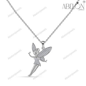 Puede incluir: Un collar plateado con un colgante de hada. El hada presenta detalles brillantes en las alas, el vestido y la cara. La cadena es fina, y la marca "ABELON JEWELS" es visible. Una joya encantadora.