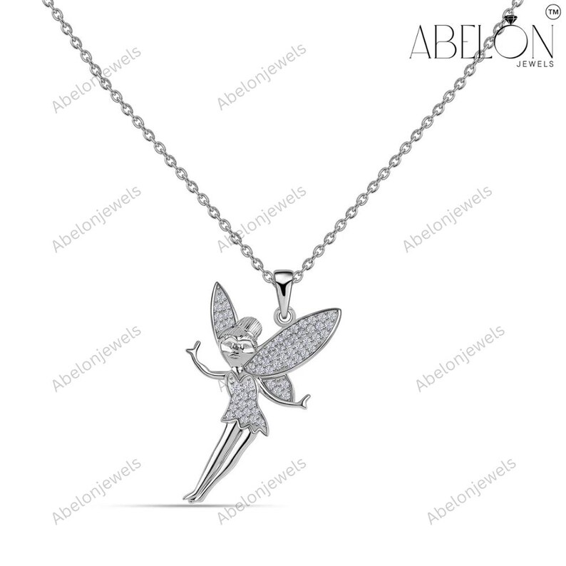 Puede incluir: Un collar plateado con un colgante de hada. El hada presenta detalles brillantes en las alas, el vestido y la cara. La cadena es fina, y la marca "ABELON JEWELS" es visible. Una joya encantadora.