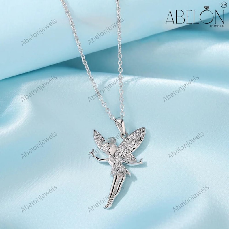 Puede incluir: Un collar plateado con un colgante de hada. El hada tiene los brazos extendidos y alas brillantes. El colgante est&aacute; detallado con peque&ntilde;as piedras transparentes. El collar se muestra sobre una tela de sat&eacute;n azul claro. La marca "ABELON JEWELS" es visible.