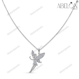 Puede incluir: Un collar de plata con un colgante de hada. El hada tiene los brazos extendidos y alas detalladas, todas incrustadas con peque&ntilde;as piedras brillantes. La cadena del collar es delicada y el colgante est&aacute; centrado. La marca "ABELON JEWELS" es visible.