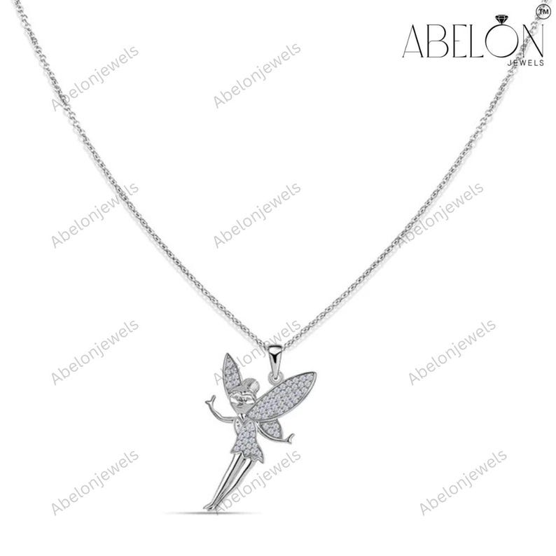Puede incluir: Un collar de plata con un colgante de hada. El hada tiene los brazos extendidos y alas detalladas, todas incrustadas con peque&ntilde;as piedras brillantes. La cadena del collar es delicada y el colgante est&aacute; centrado. La marca "ABELON JEWELS" es visible.