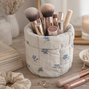 Könnte beinhalten: Ein weißer, gesteppter Make-up-Pinselhalter mit blauem Blumenmuster, gefüllt mit verschiedenen Make-up-Pinseln und Kosmetikprodukten. Der Halter steht auf einer Holzoberfläche, zusammen mit einem Haargummi, Ringen und einer Kerze.