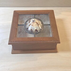 May include: A square wooden box with a hinged lid featuring a central circular seashell inlay surrounded by textured, translucent glass panels held in place by dark metal framing. The box has a smooth, medium-brown finish on all visible sides.
