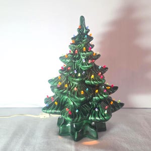 May include: A dark green ceramic Christmas tree figurine with a glossy finish, featuring multiple tiers of moulded branches. The tree is illuminated by numerous small, multi-coloured plastic light bulbs in red, yellow, blue, and clear tones.