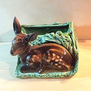 May include: A ceramic planter featuring a brown fawn with white spots resting in front of a green textured container designed to resemble a forest clearing with foliage and branches. The glossy finish highlights the deer's form and the greenery.