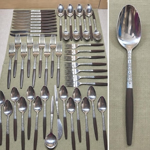 Puede incluir: Un gran juego de cubiertos de acero inoxidable con mangos de plástico marrón oscuro y un cuello de metal plateado con un intrincado patrón de volutas. La colección incluye varios cuchillos, tenedores y cucharas en filas.