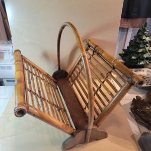 Puede incluir: Un revistero vintage de bambú con asa. El revistero está hecho de bambú marrón claro y tiene forma rectangular con asa. El revistero está abierto por ambos lados y tiene una base de madera. Está diseñado para guardar revistas o periódicos.