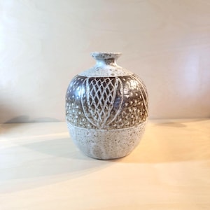 以下が含まれることがあります： 狭い首を持つ丸い陶器製の花瓶。クリーム色の斑点があるベースと、濃い茶色の上部で構成されています。表面全体には、アザミのような様式化された形状と小さな点が繰り返されるエッチング加工の植物模様が施されています。