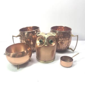 May include: A collection of copper-toned items including two hammered mugs with handles, a small three-legged bowl, a central owl-shaped container with green eyes, and a tiny handled scoop, all arranged together on a plain white surface.