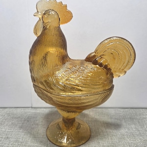 Puede incluir: Un plato de vidrio color ámbar translúcido con forma de gallo y tapa. La pieza presenta una textura moldeada detallada que representa plumas en el cuerpo, las alas y la cola, y se apoya sobre una base circular con una superficie texturizada.