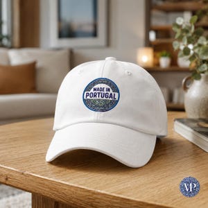 May include: White baseball cap with a curved brim. The cap features a circular patch with the text "MADE IN PORTUGAL" in a blue and white design. The cap is sitting on a wooden table.