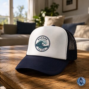 May include: A trucker hat with a white front panel and a navy blue brim. The hat features a circular emblem with the word "NAZARE" above a graphic of a large wave. The words "BIG WAVE" are below the wave graphic. The hat has a black mesh back.