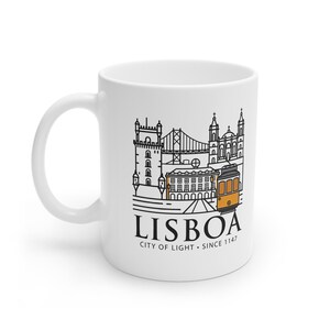 May include: White ceramic mug featuring a black and orange line drawing of Lisbon landmarks, including a bridge, buildings, and a tram. The word "LISBOA" is printed below the illustration, along with "CITY OF LIGHT. SINCE 1147."